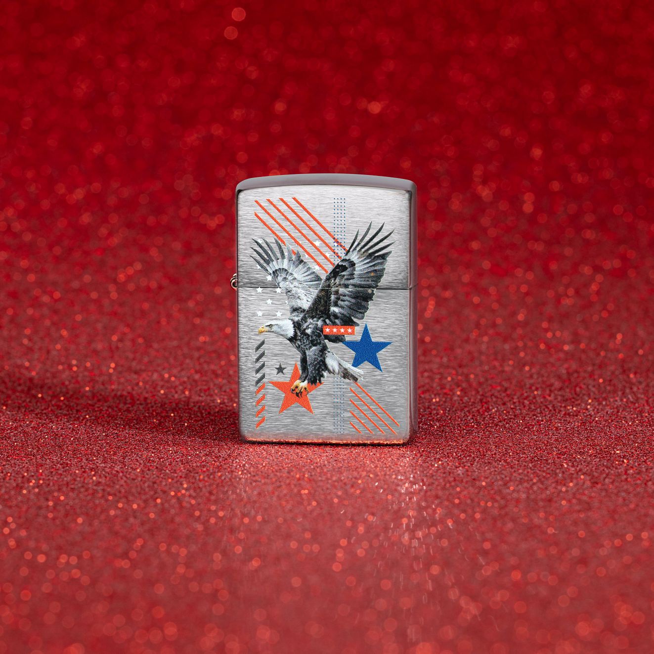 Lifestyle image of Zippo Abstract Americana Design Brushed Chrome Windproof Lighter standing with a red glitter background showing the front of the lighter. 


