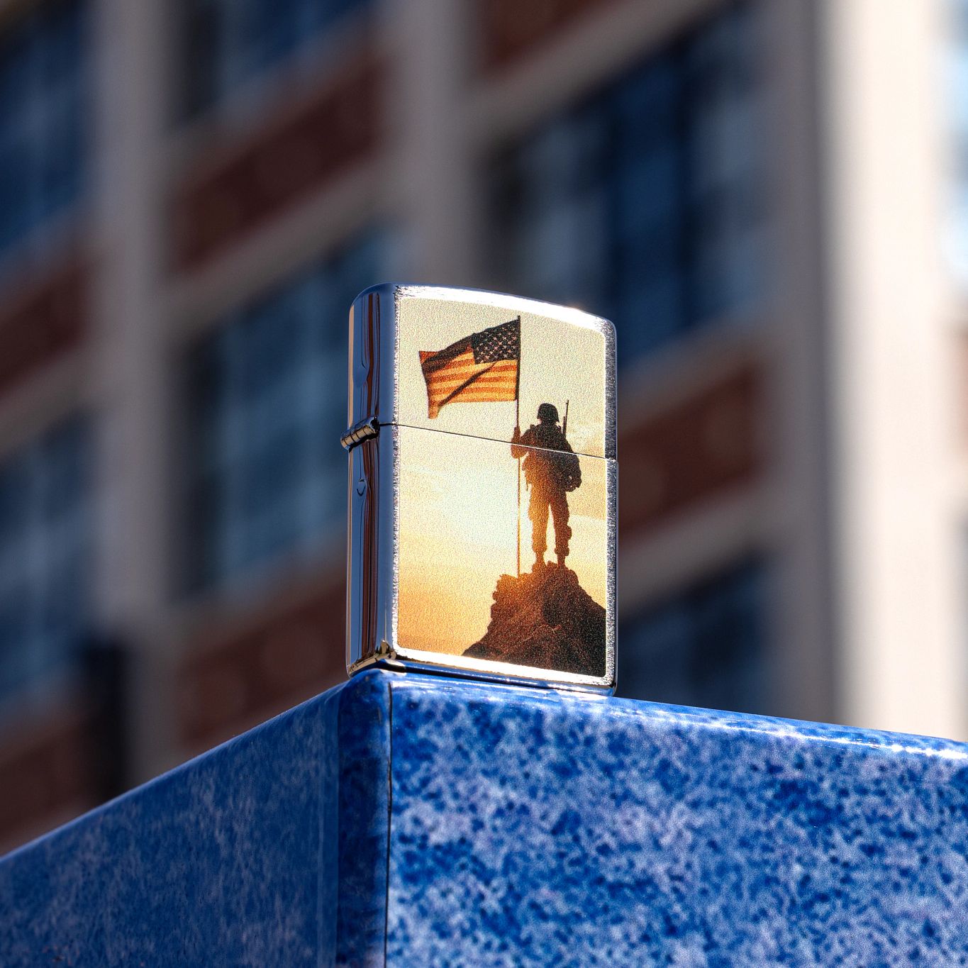 Lifestyle image of Zippo Soldier and Flag Design Brushed Chrome Windproof Lighter standing on the corner of a bright blue stone wall, showing the front and hinge side of the lighter with a blurred building as the background. 

