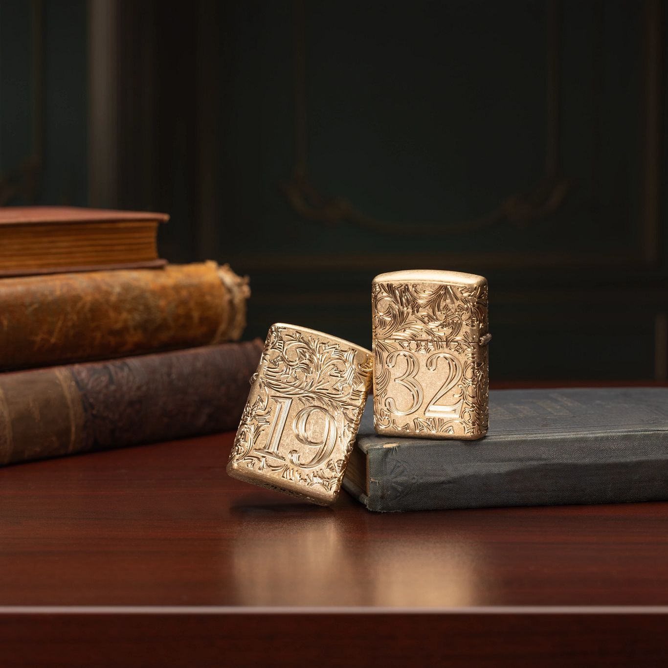 Lifestyle image of two Zippo 1932 Filigree Design Armor® Tumbled Brass Windproof Lighters; one showing the front of the lighter and the other showing the back and hinge side of the lighter, both standing on a wooden desk with four books and a dark background. 

