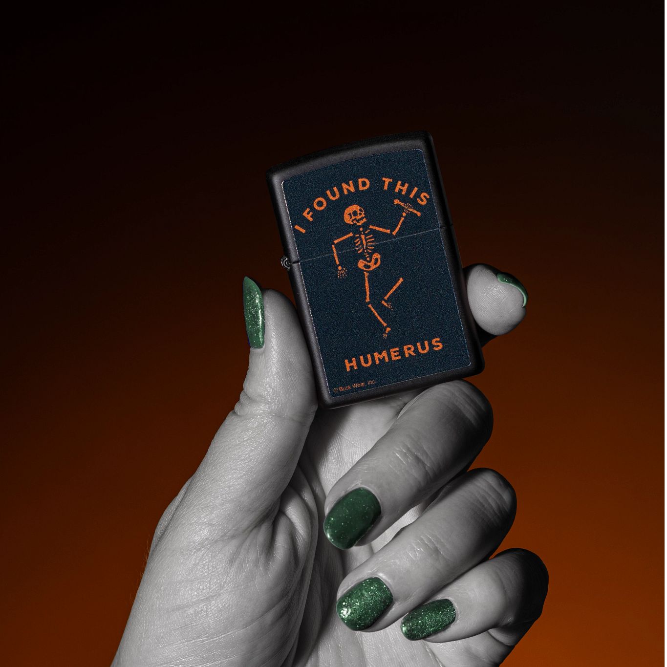 Lifestyle image of Zippo Buck Wear™ Humerus Design Black Matte Windproof Lighter being held in a hand with a black and orange ombre background. 

